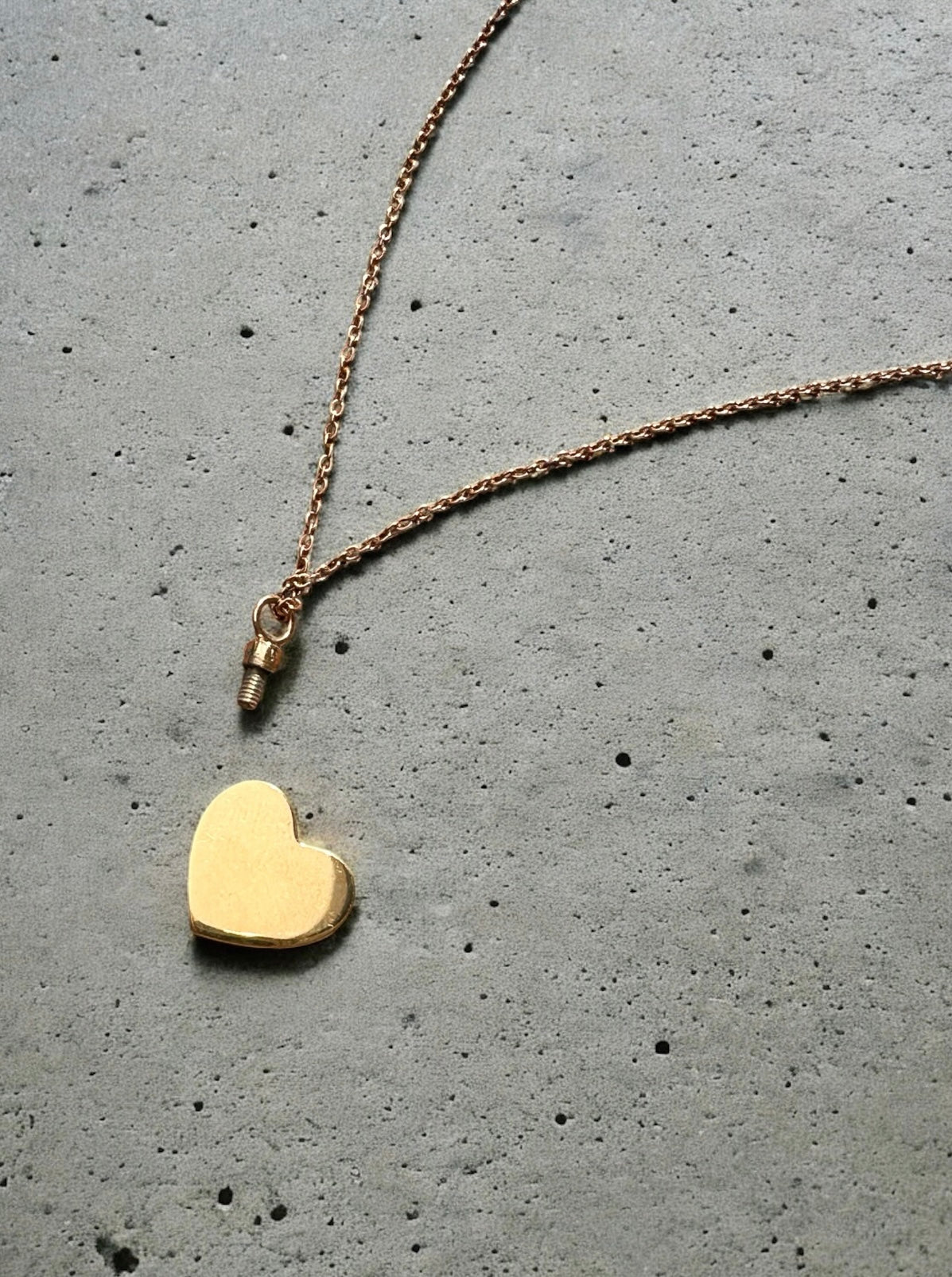 Gold heart-shaped pendant shown open to reveal screw closure and hollow interior on a chain against a gray concrete background