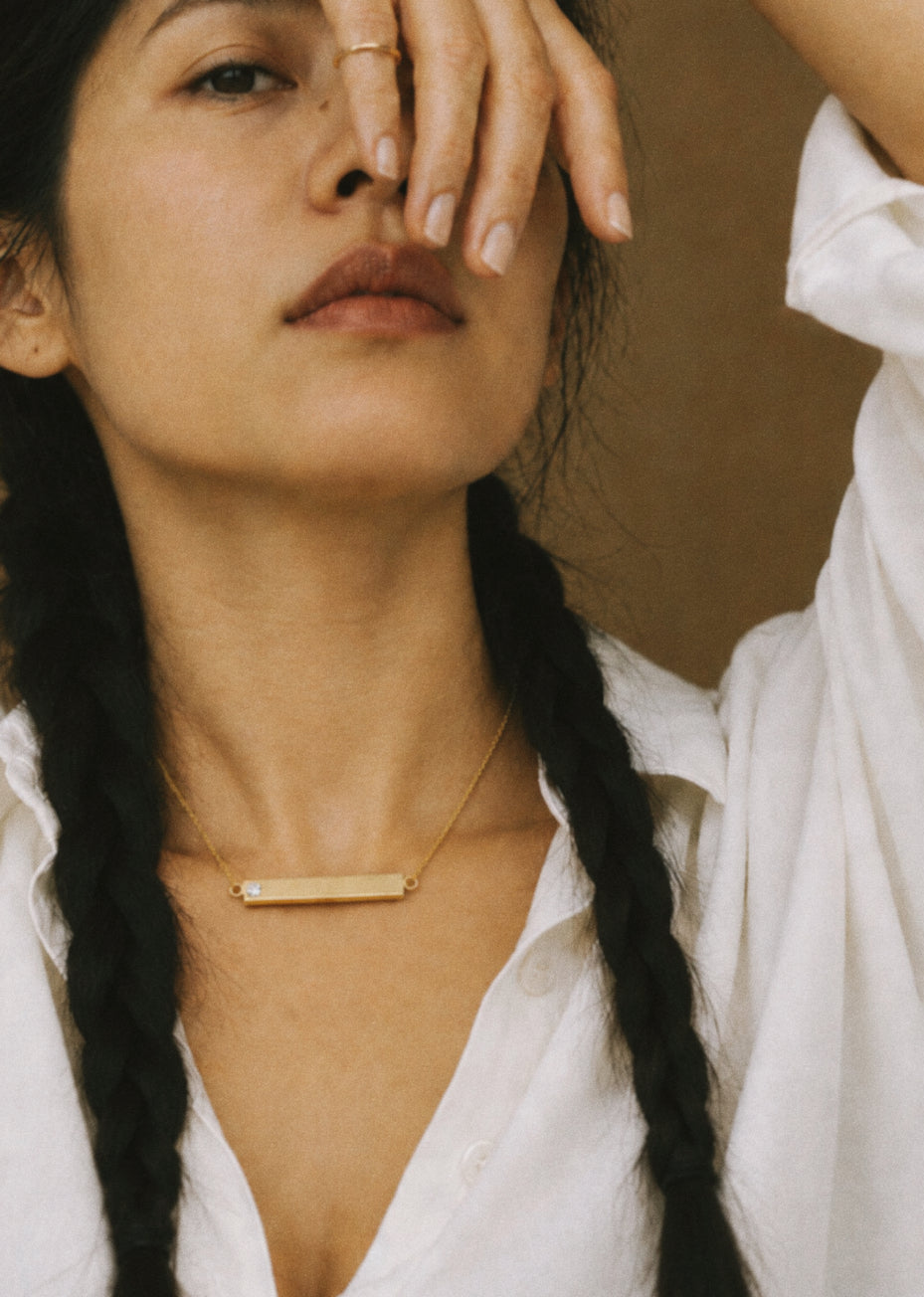 A close-up lifestyle shot of a woman with dark braided hair wearing a delicate gold bar memorial necklace with hollow interior, designed to hold cremation ashes