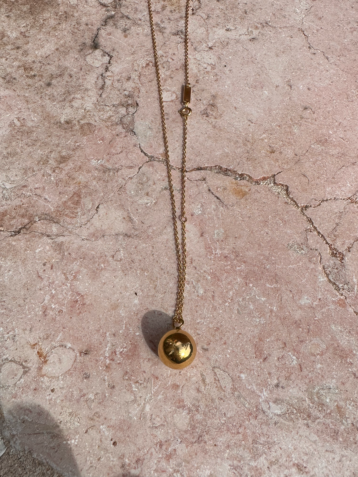 Gold necklace with a sphere urn pendant with hollow interior to hold ashes on a textured pink surface