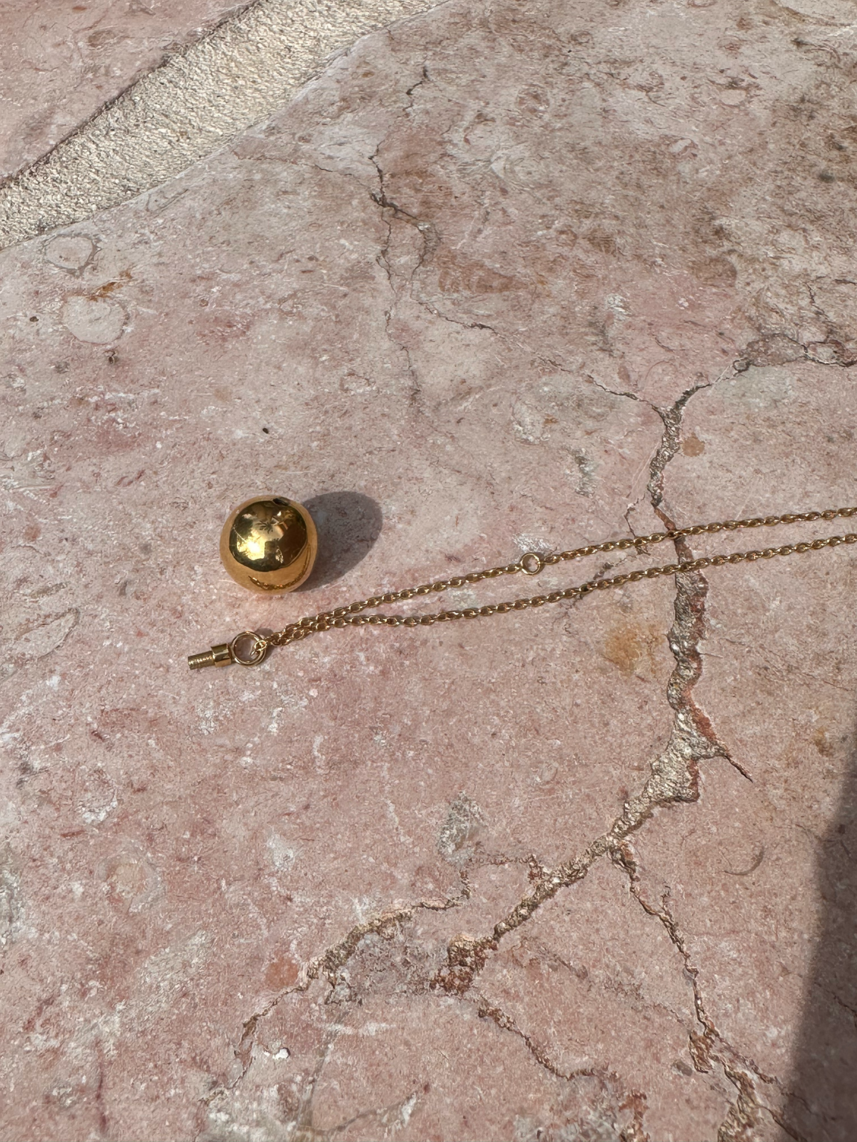 Gold necklace with a spherical pendant shown open with screw closure, hollow interior to hold memorial ashes on a textured pink surface