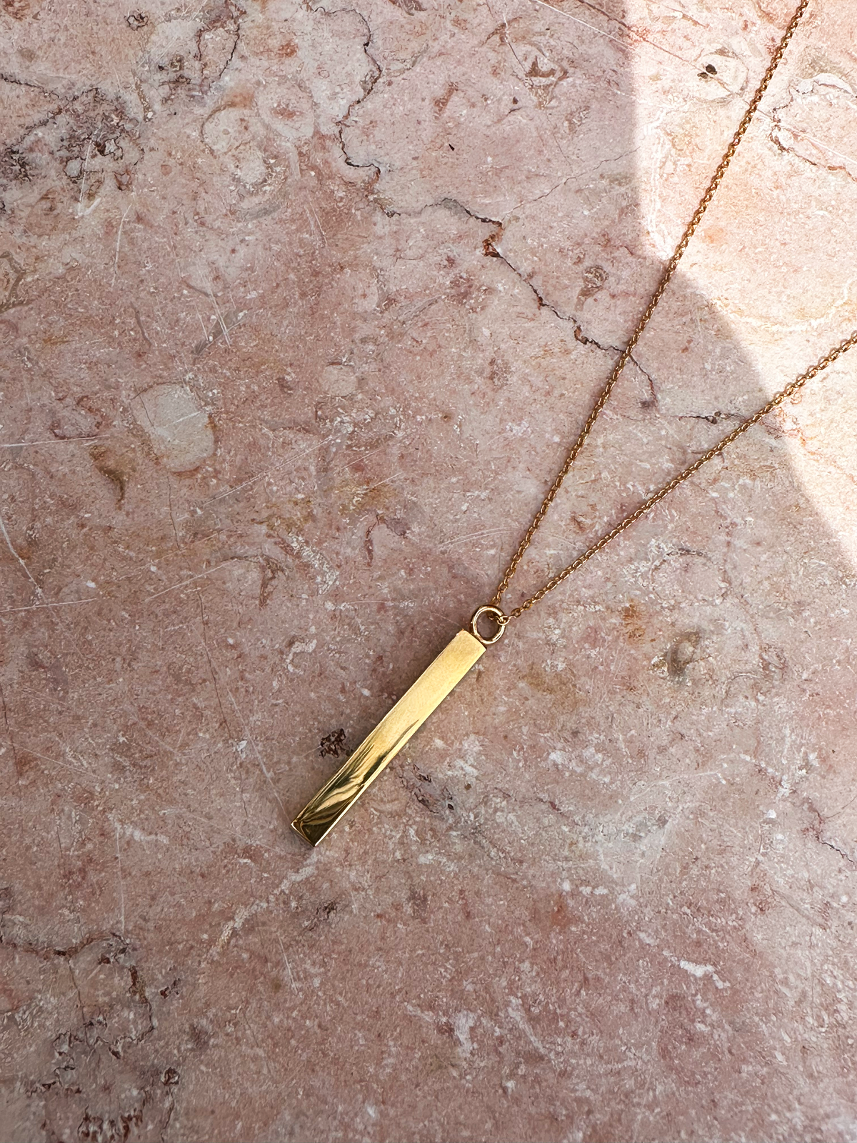 Gold urn necklace with a hollow bar pendant to hold ashes on a pink marble surface