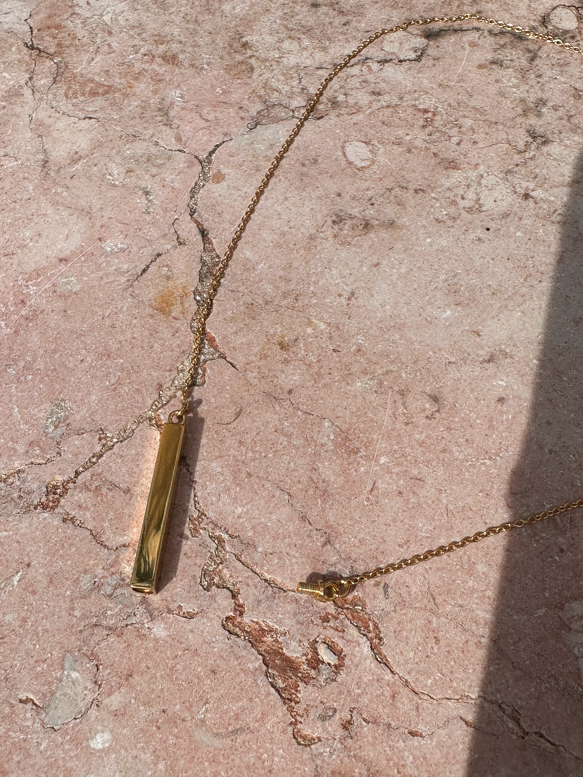 Gold necklace with a bar pendant, shown open with a screw enclosure to hold cremation ashes, on a textured stone surface
